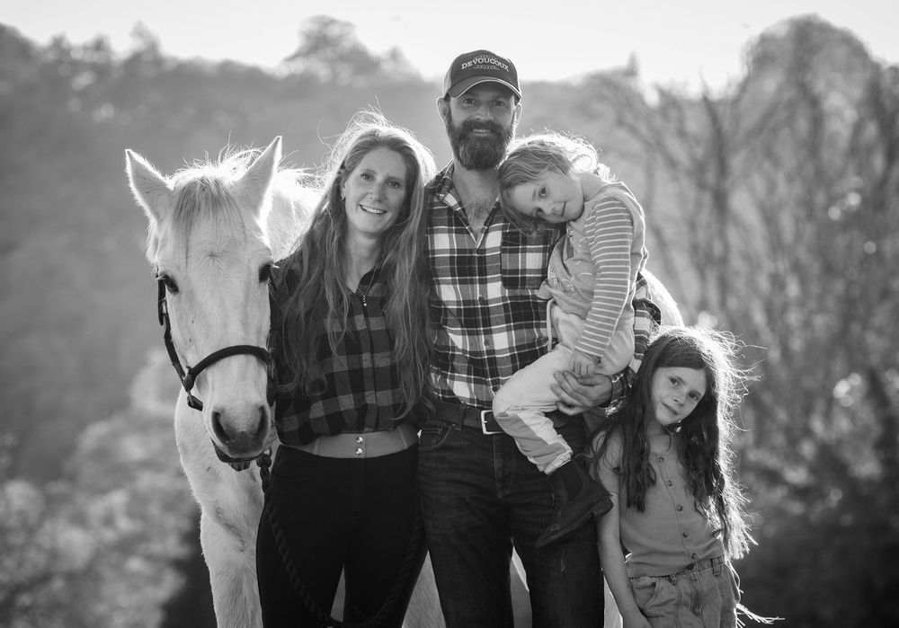 Black and white family portrait series with white horse in rustic outdoor setting.