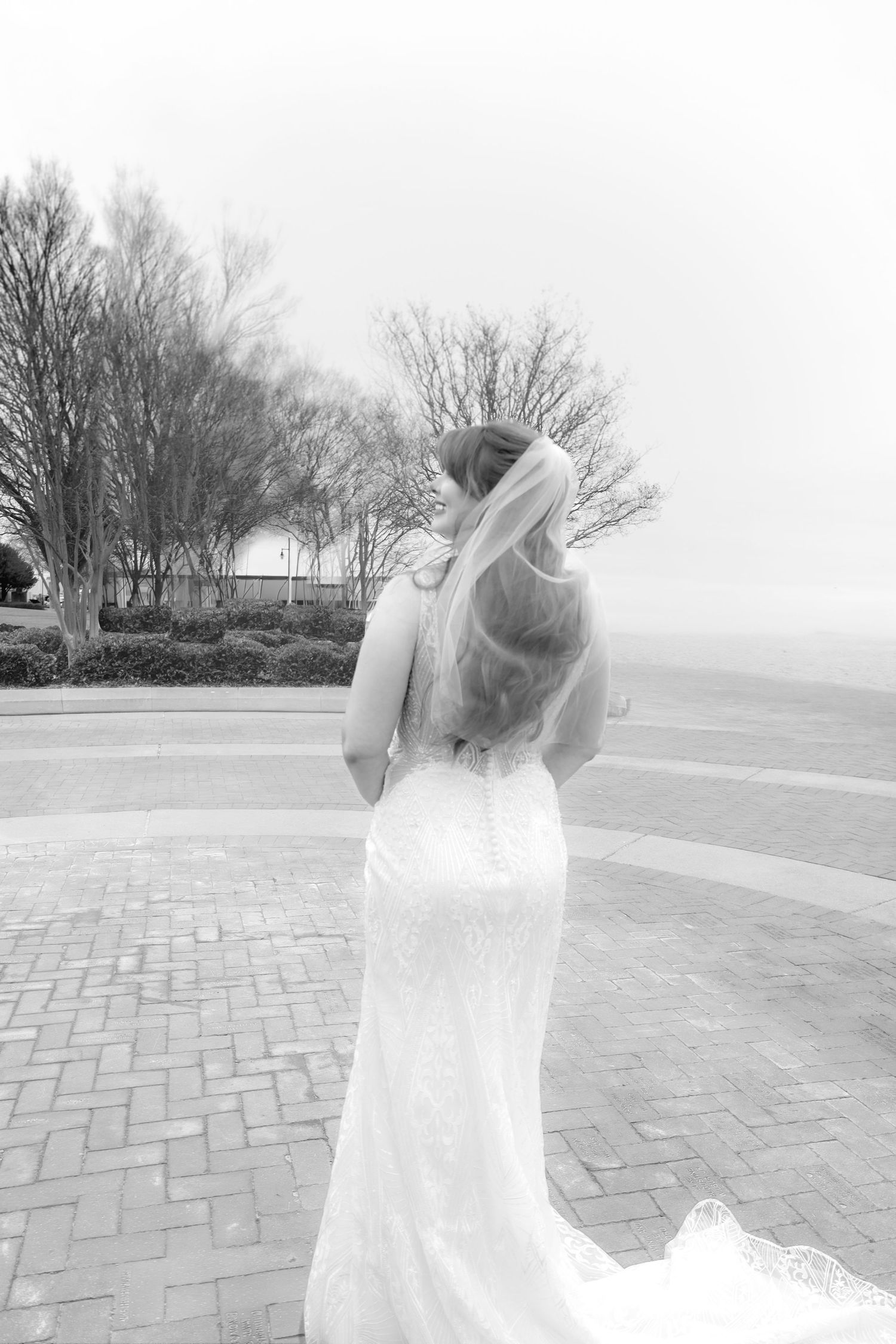 Black and white photo of a bride in a fitted lace gown with veil looking over her shoulder outdoors.