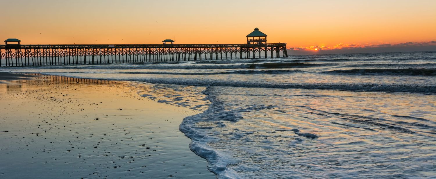 Panoramic view of a wooden fishing pier stretching into the ocean at sunset with golden sky and rolling waves.