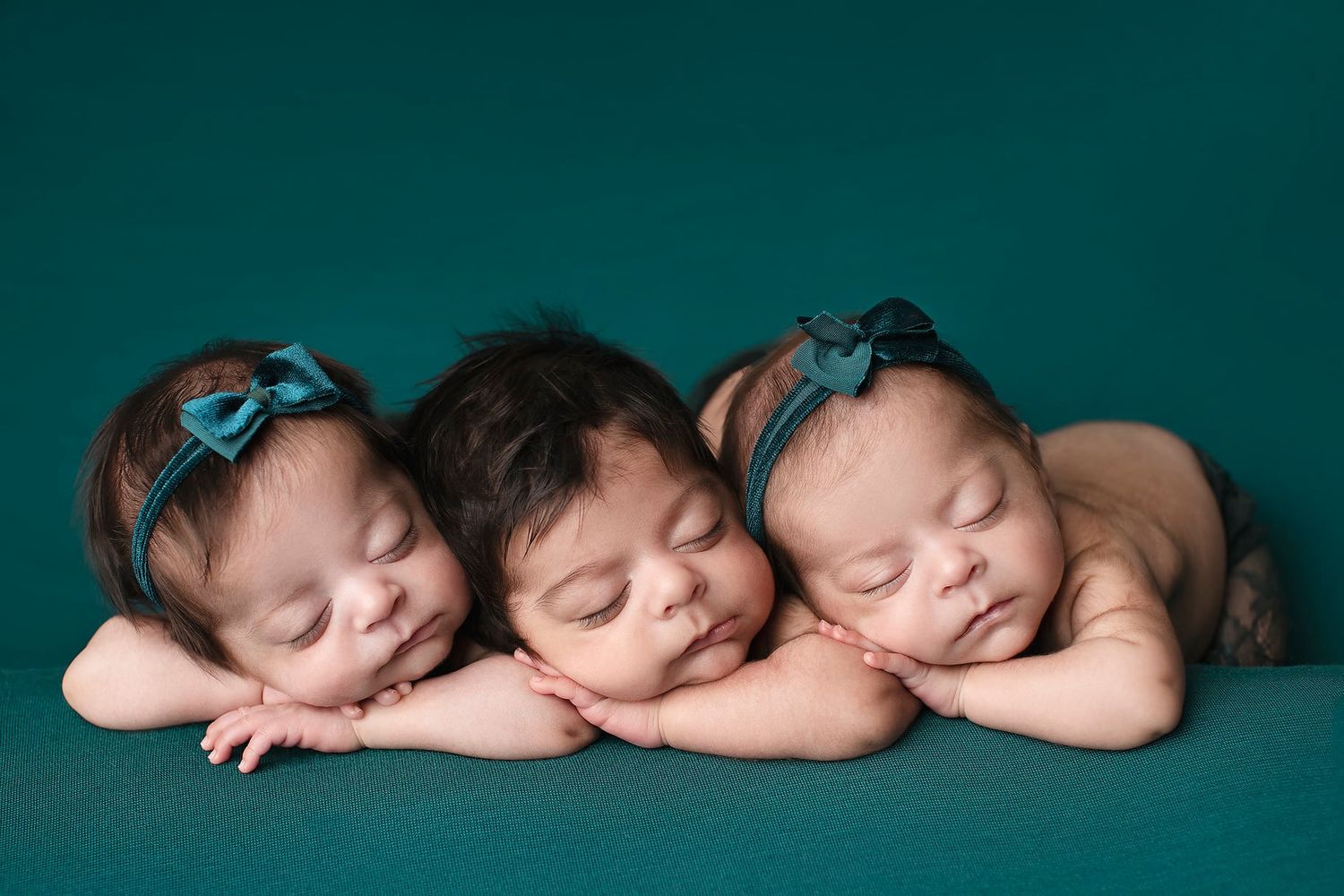 Three sleeping newborn babies wearing matching blue bows lie in a row on a dark background. Elizabeth Lang Photography Surrey