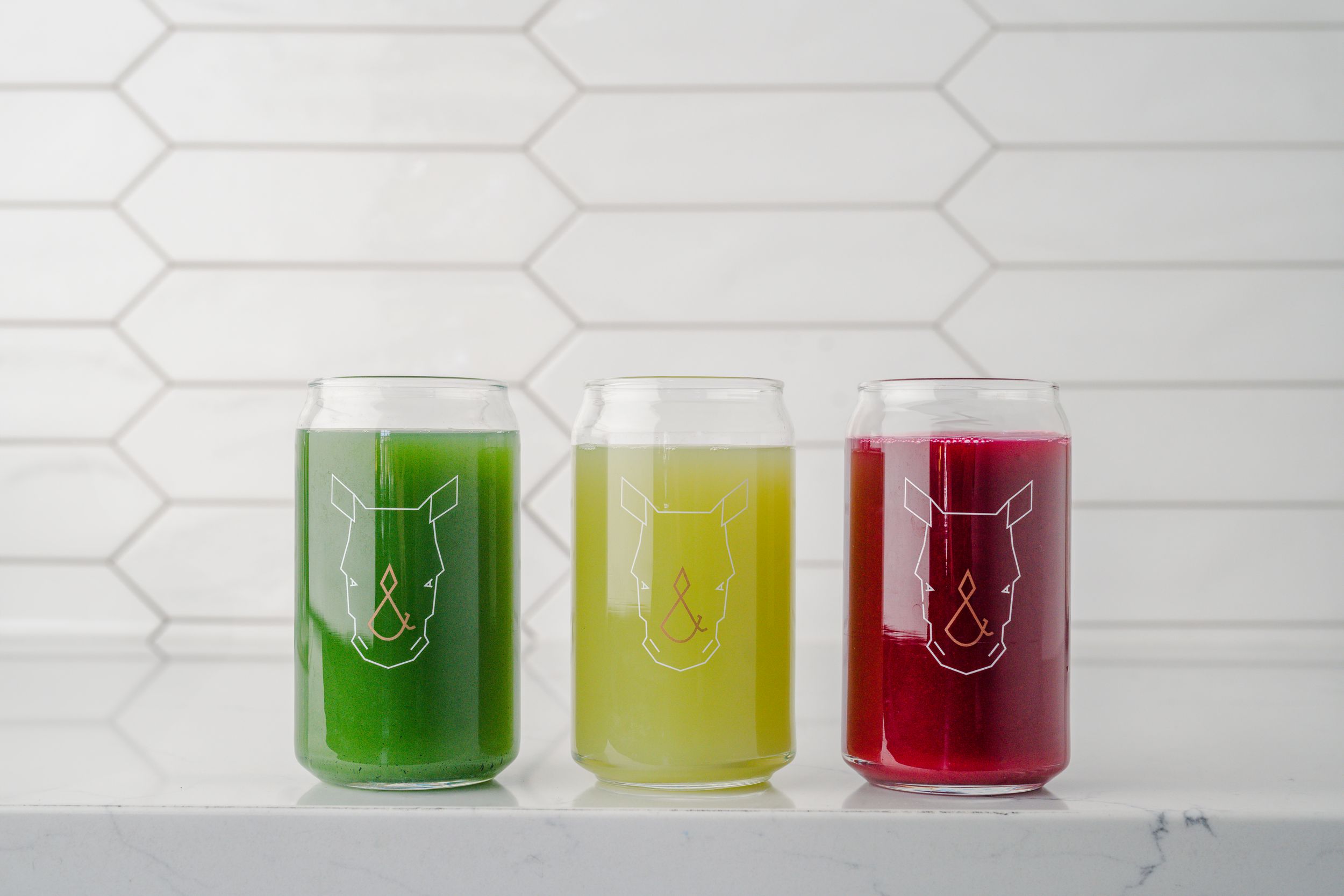 Three vibrant glasses containing green, yellow, and red juice drinks lined up against a white hexagonal tile wall.