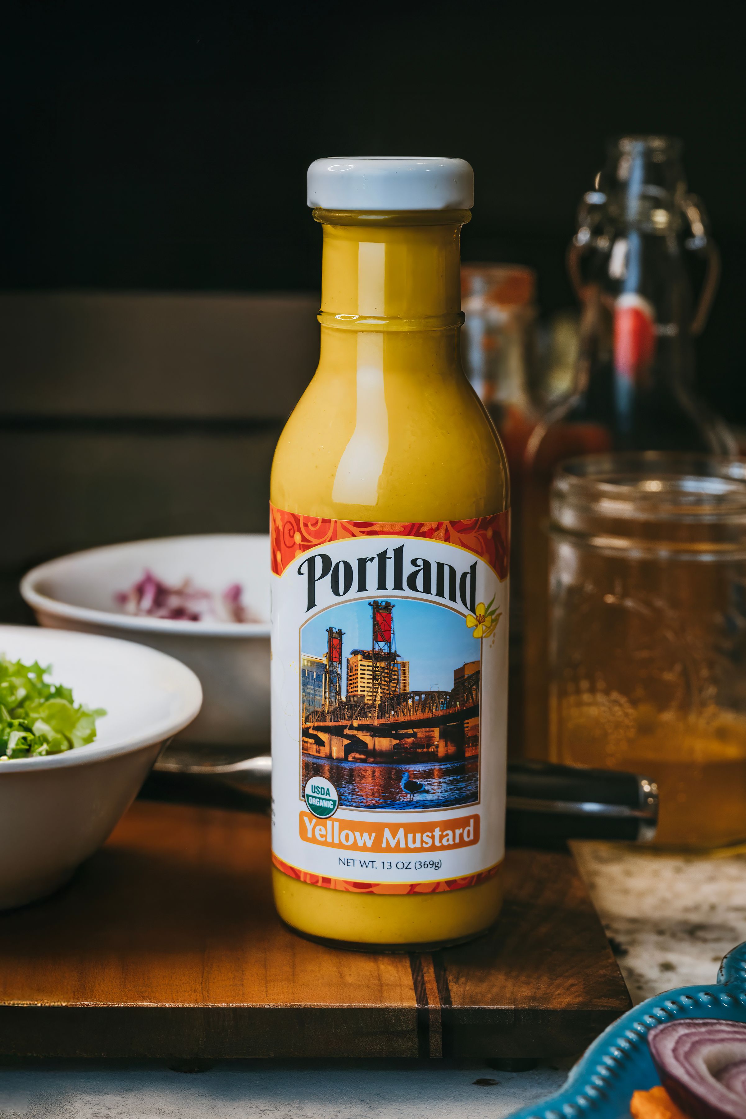 A bottle of Portland yellow mustard with a cityscape label sits on a wooden table beside bowls of food and a glass.