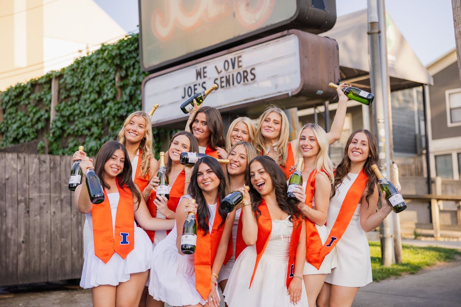 Group of UIUC girls in white dresses in front of Joe's Brewery holding champagne bottles