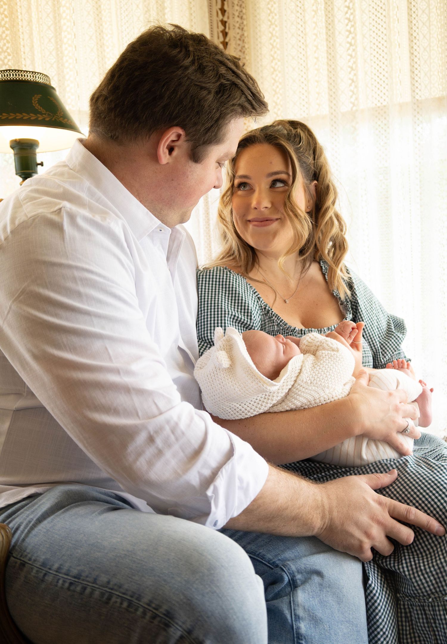 Happy couple in matching green gingham outfits share tender moment with newborn baby in floral wallpaper room.