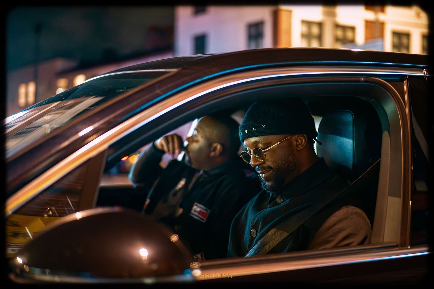 A brown car drives through a city street at night with illuminated buildings visible in the background.