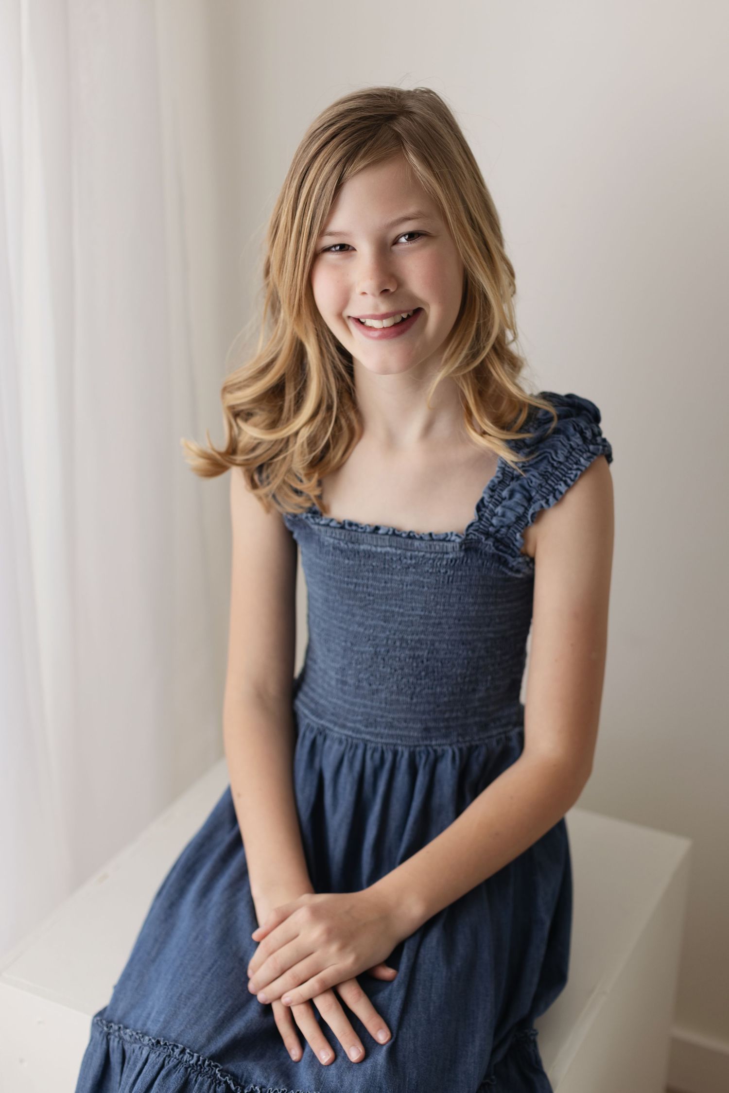 Child in a navy blue smocked dress poses against a white wall in a series of playful portraits. Elizabeth Lang Photography Surrey