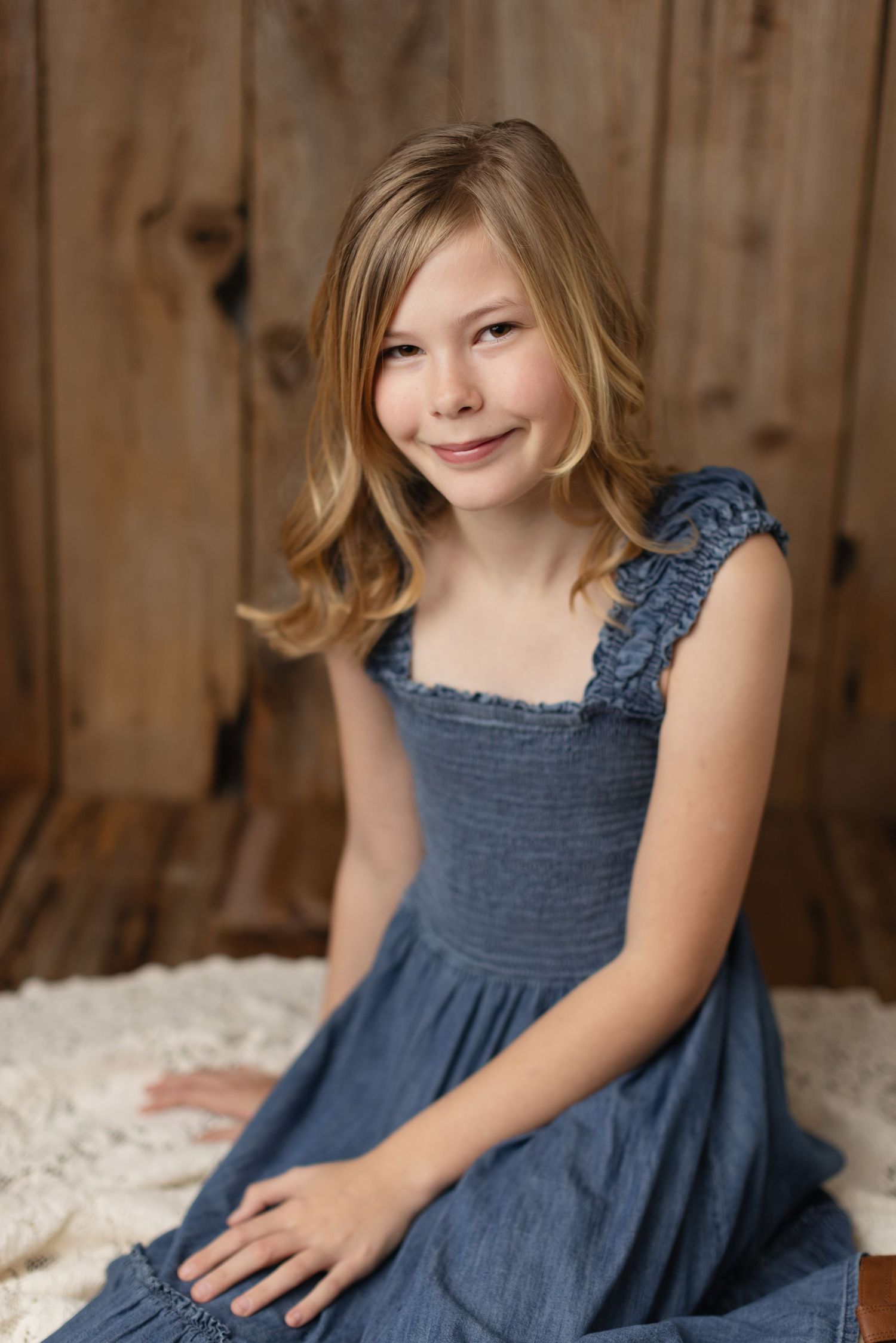 A young child in a navy blue dress sits on a white rug against a rustic wooden wall background. Elizabeth Lang Photography Surrey