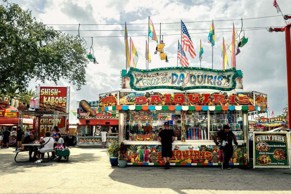 Colorful carnival food stand with flags and signage at a fairground under cloudy skies.