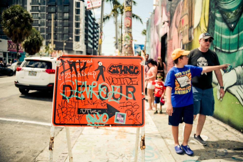 Orange detour sign with graffiti art on urban street with buildings and murals.