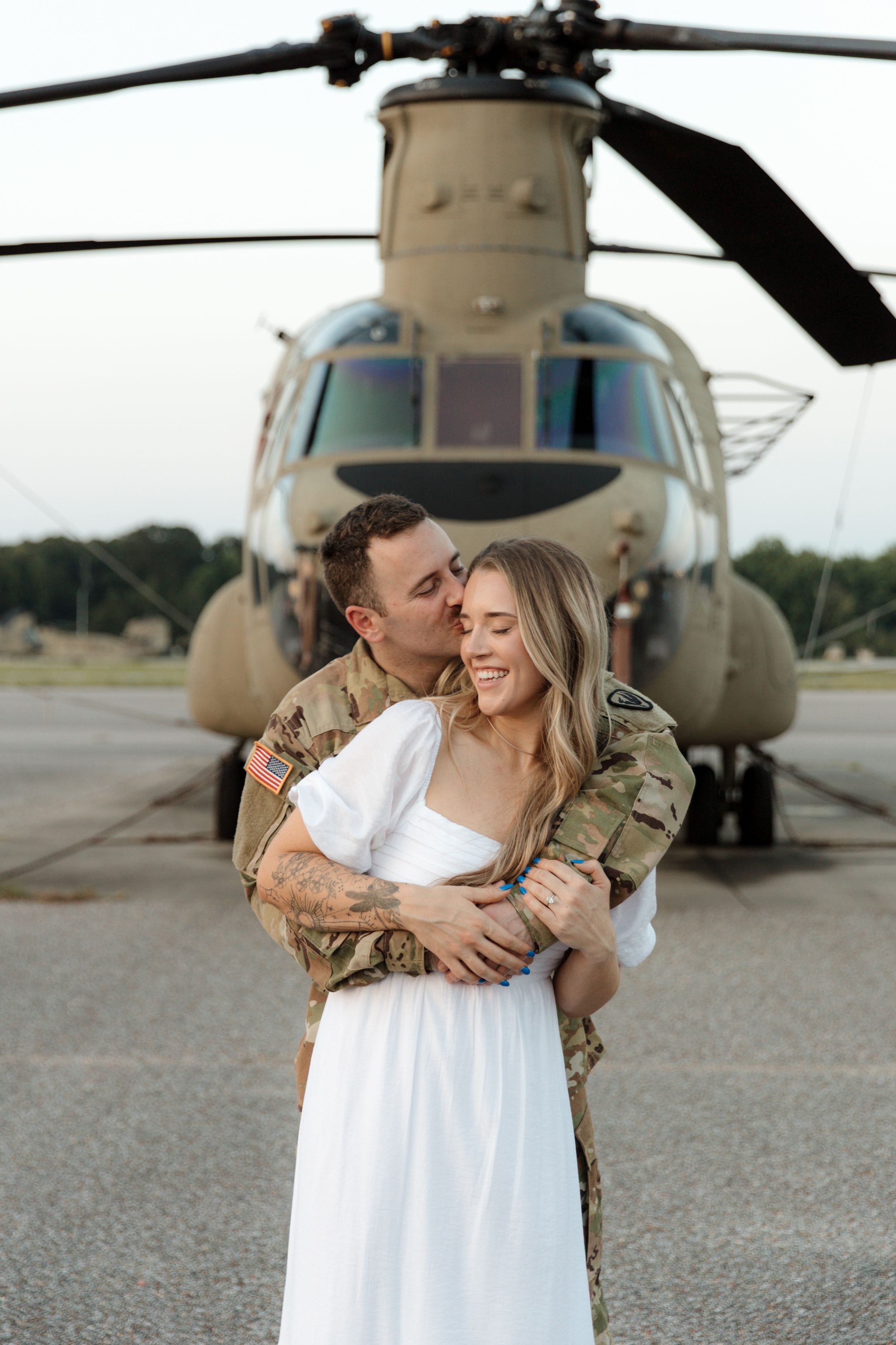 A tender moment between a couple in front of a military helicopter on an airfield.