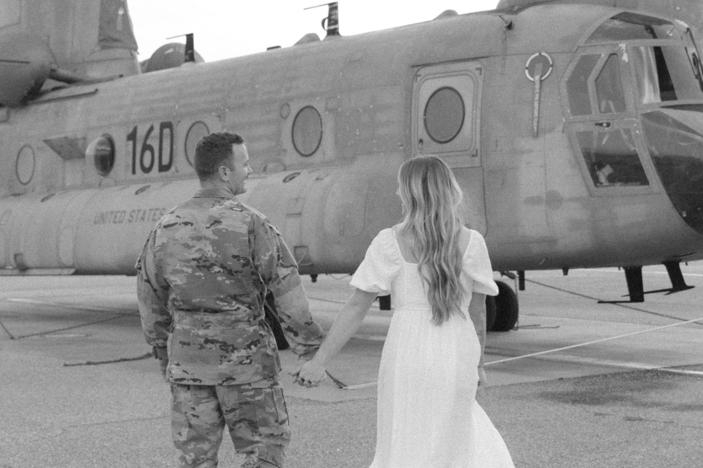 A couple walks hand in hand toward a military helicopter on an airfield.
