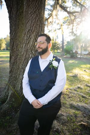Wedding portrait of person in navy vest sitting against tree in natural sunlight.