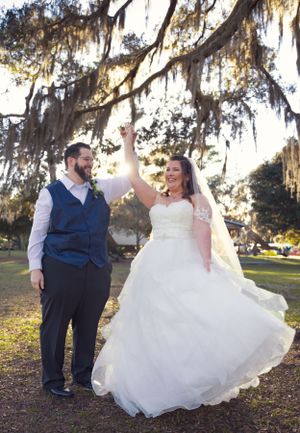 A joyful wedding couple dancing and twirling under moss-covered trees in a park setting.