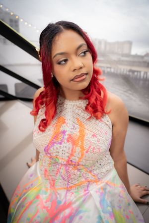 Person in white beaded dress with vibrant splatter details and red curls poses near window.