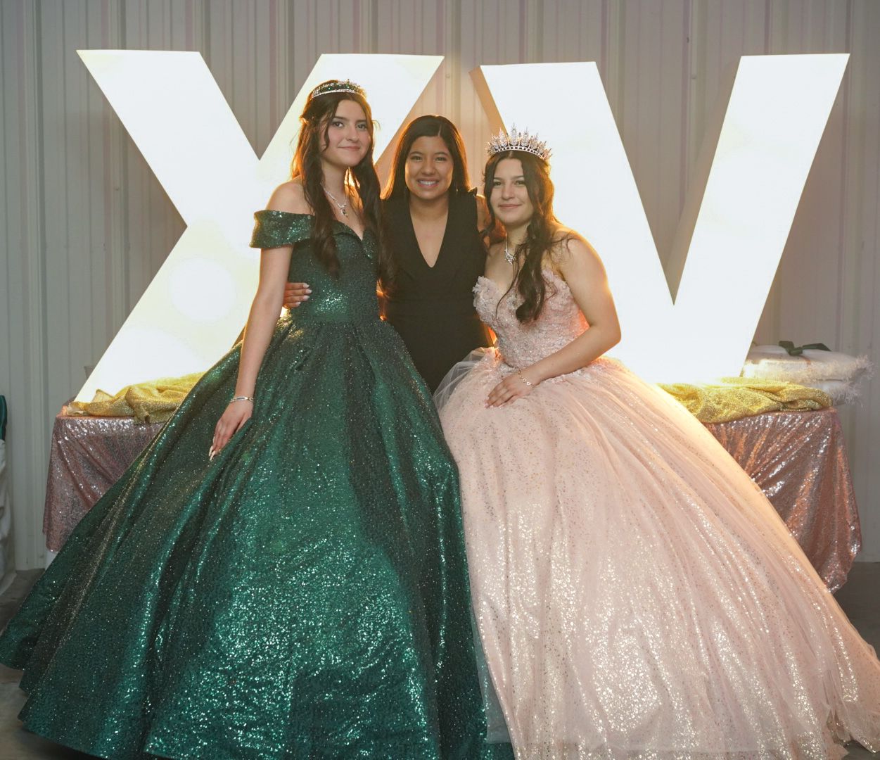 Three people pose together at a formal event, wearing elegant gowns in emerald green and blush pink against a white backdrop.