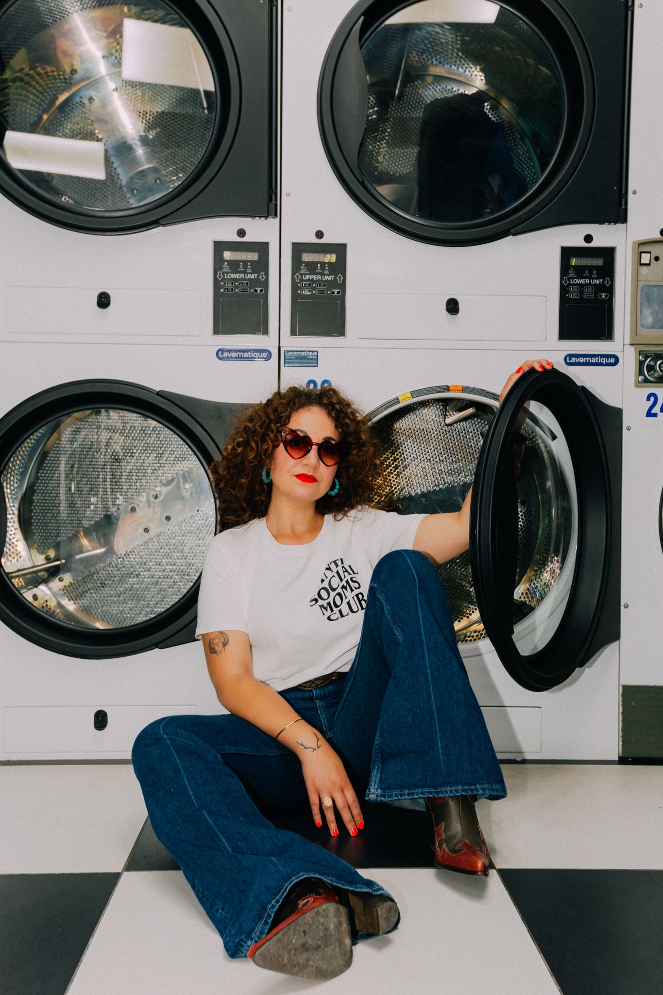 Curly-haired woman in white t-shirt and flared jeans sitting on laundromat floor with washing machines behind her.