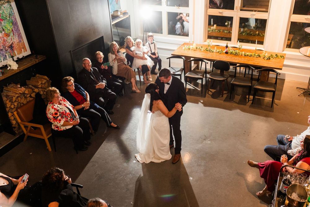 Wedding reception guests mingle and dine in a rustic indoor venue with warm lighting and wooden tables.