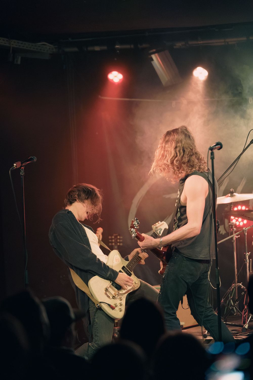 Metal band musicians perform energetically on a dimly lit stage with red lighting and silhouetted crowd in foreground.