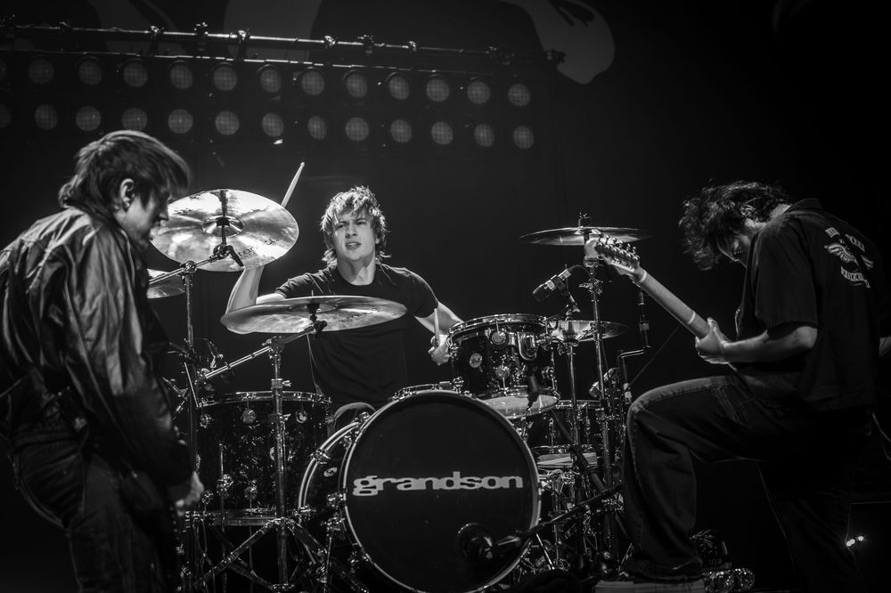 A drummer performs on stage with dramatic lighting and silhouetted figures in a live concert setting.