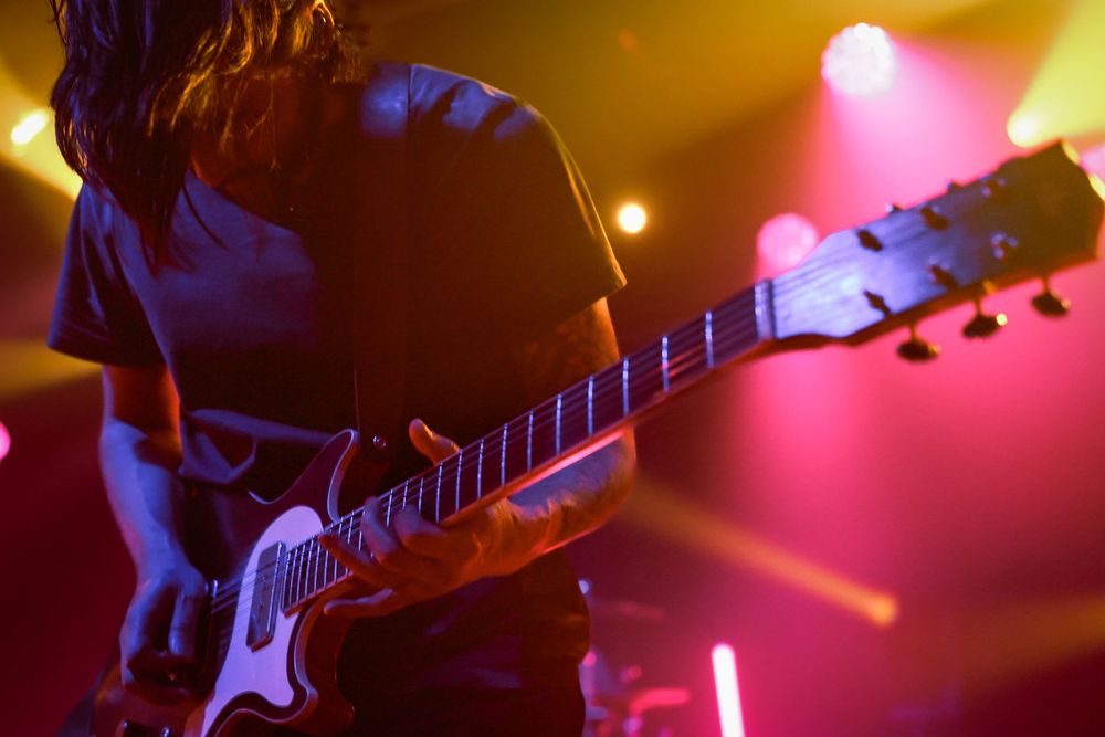 Guitarist performs on stage with colorful red and blue lighting effects creating a dramatic concert atmosphere.