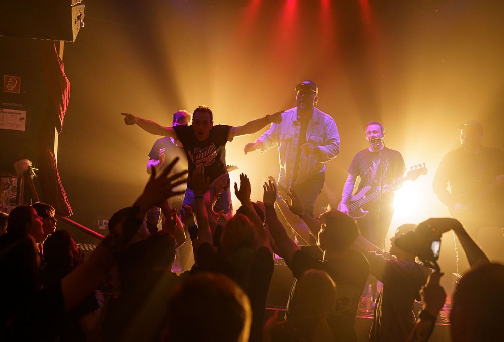 Silhouettes of performers and crowd against warm orange stage lighting during an energetic concert.
