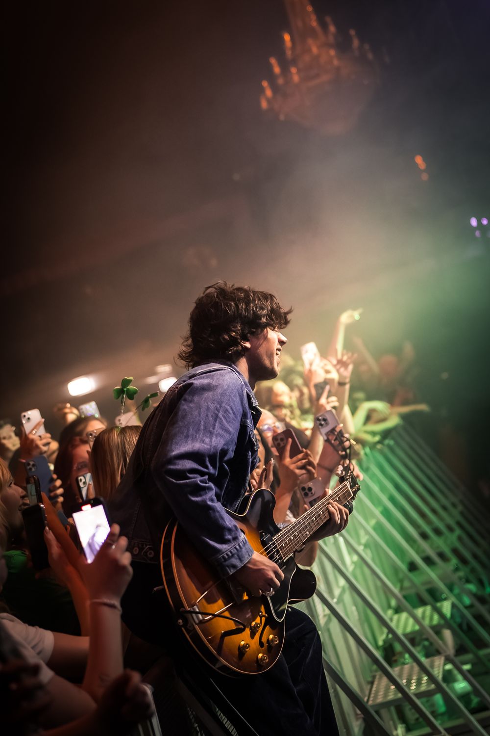 Guitarist performs on stage with dramatic backlighting while crowd raises hands in silhouette.