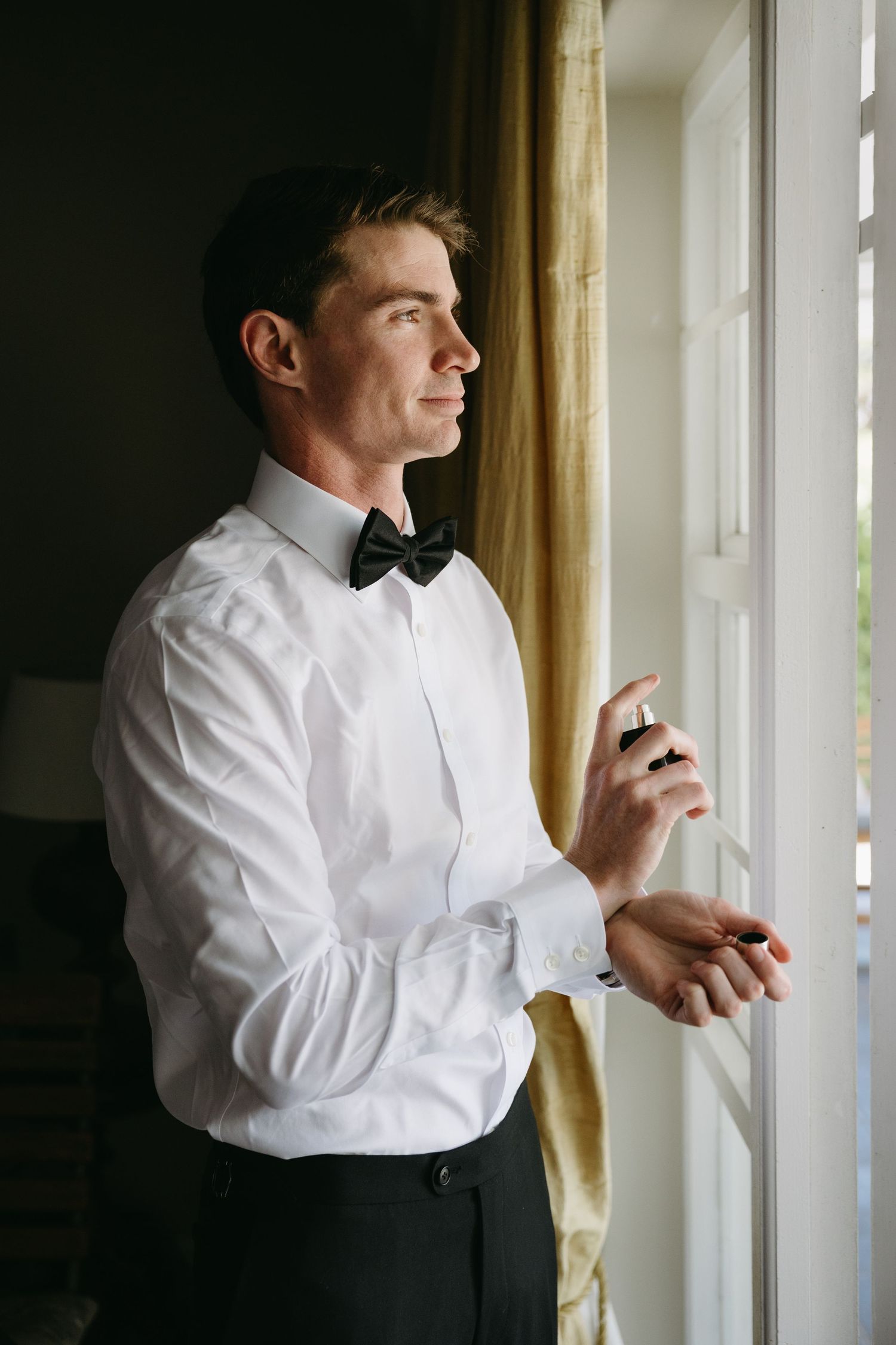 A person in formal attire adjusts their bowtie while standing near a window in moody lighting.