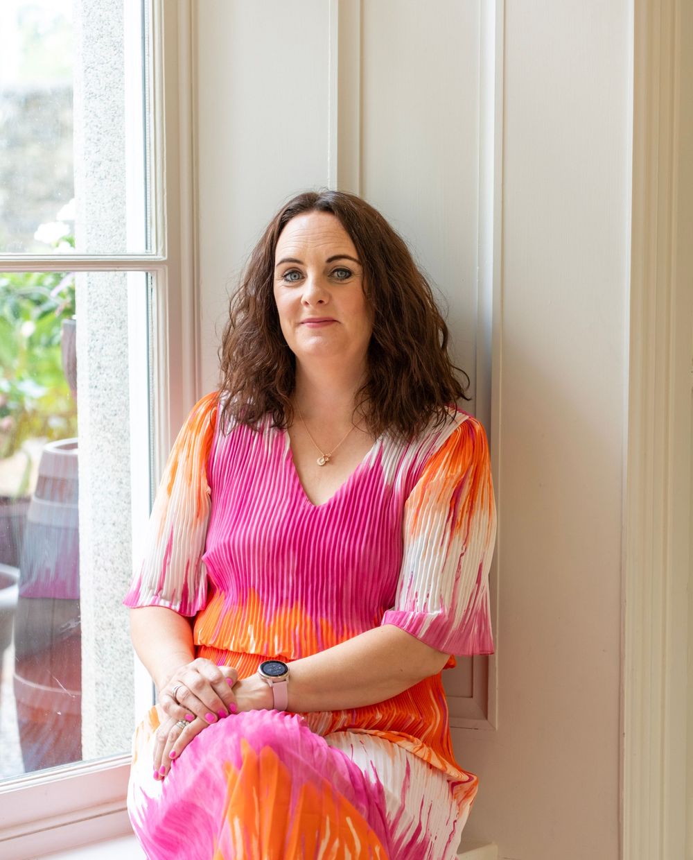 A person in a pink and orange outfit poses against a white wall near windows.