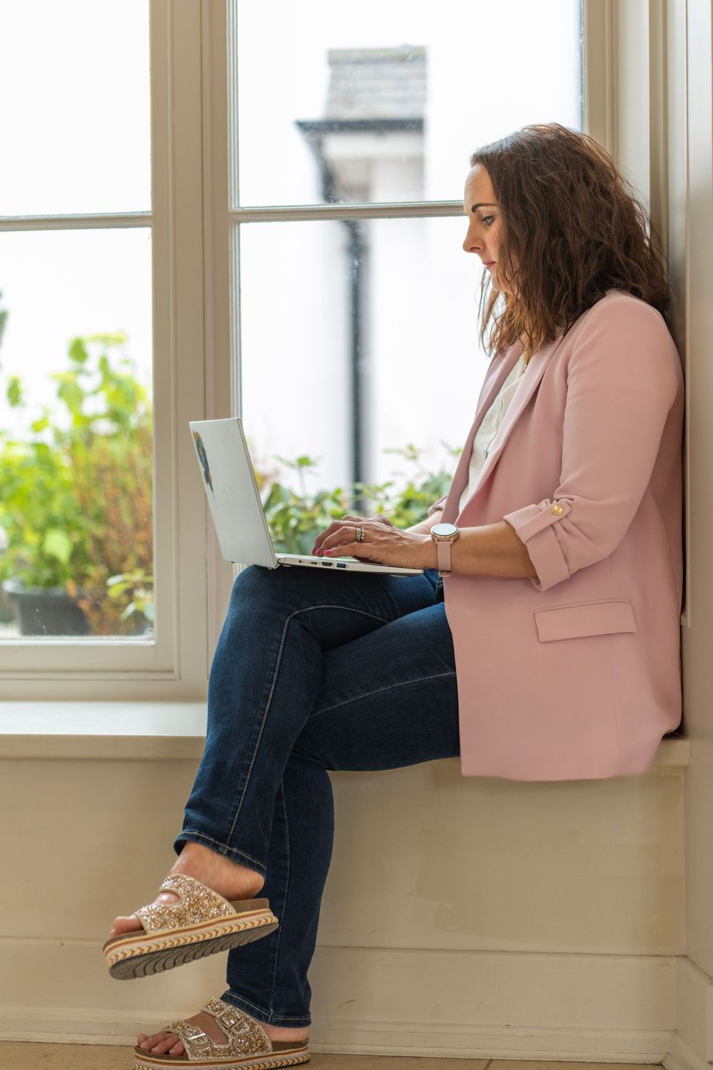 Person in pink blazer and jeans sits by window with laptop, shown from different angles in connected photos.