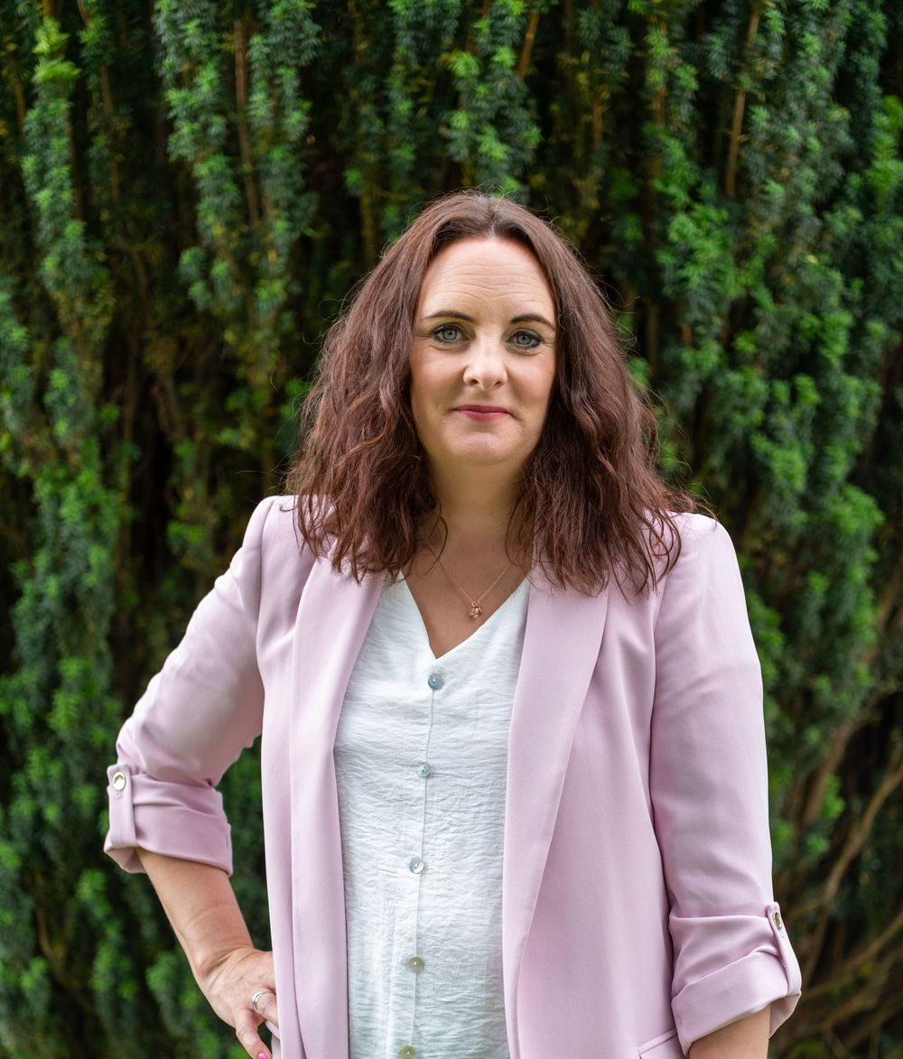 Professional portrait of someone wearing a pink blazer and white button-up shirt against a green hedge background.