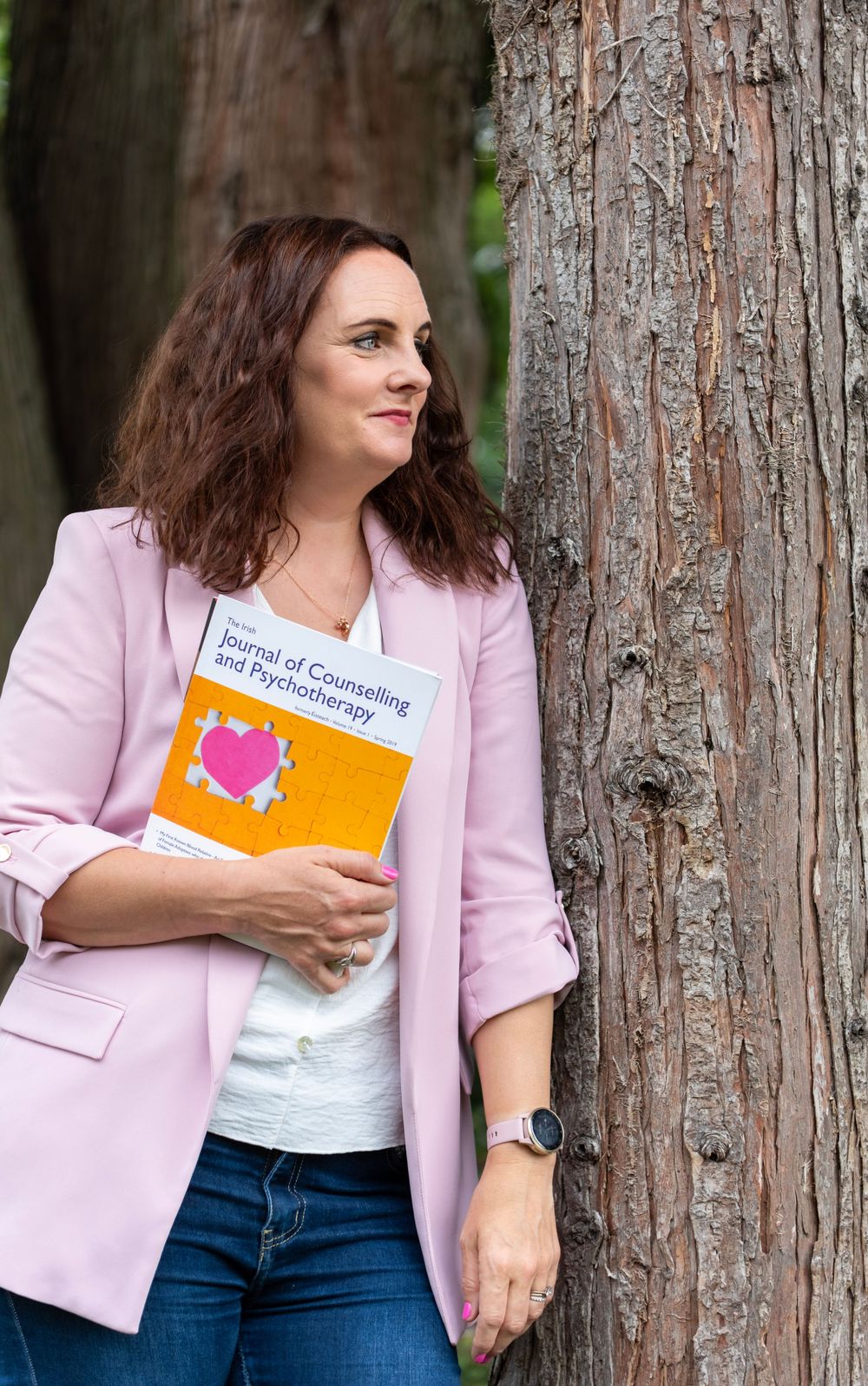 A person in a pink blazer and jeans holds a book while leaning against a tree trunk in a natural outdoor setting.