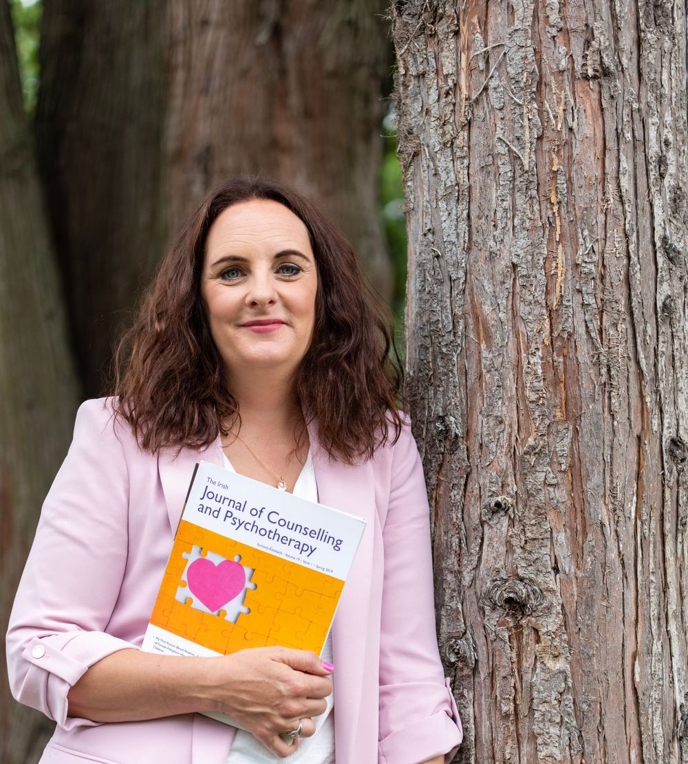 Person in pink blazer holding orange book while standing next to tree trunk in outdoor setting.