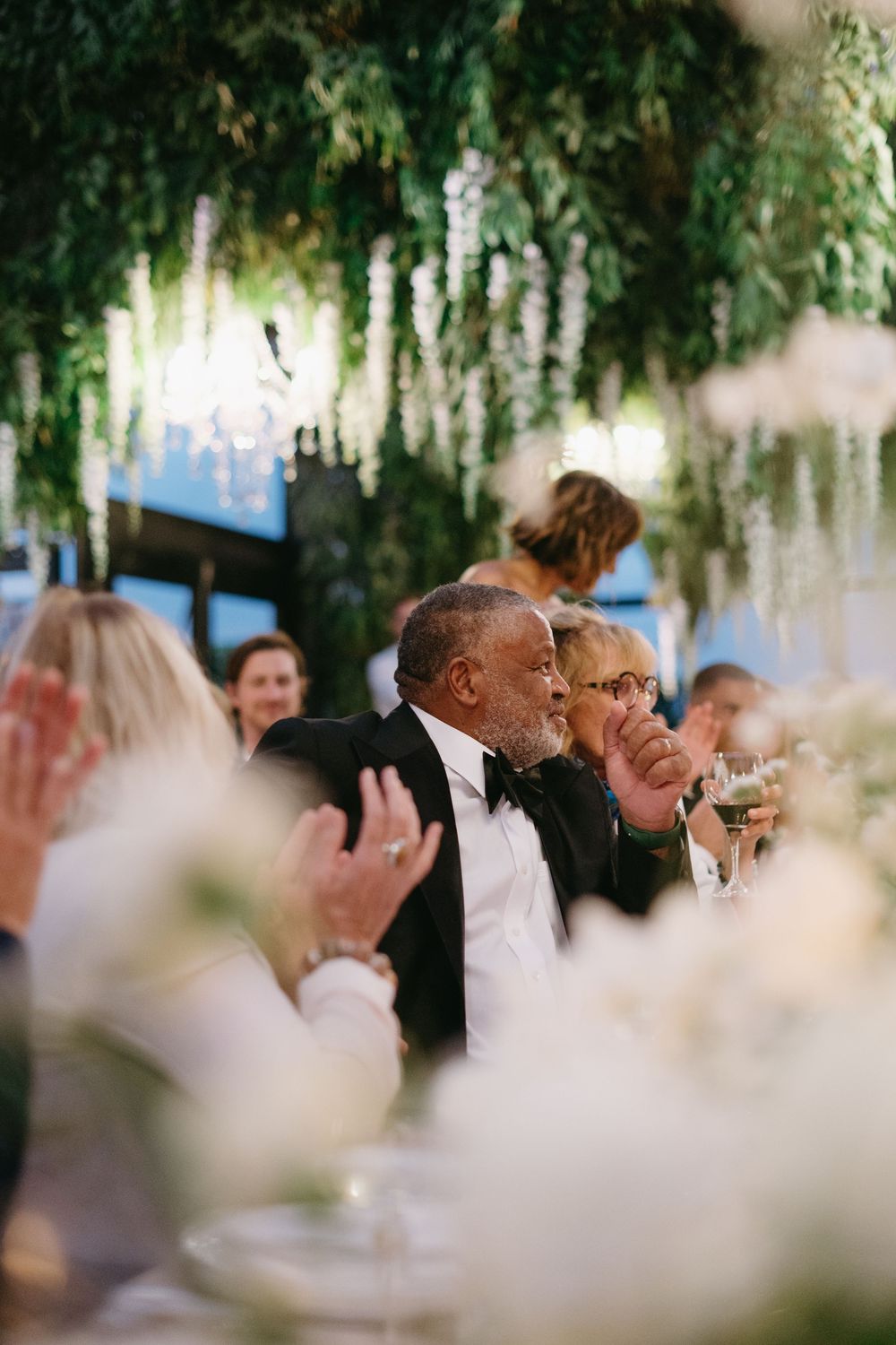 Guests enjoying an elegant outdoor wedding reception under hanging floral decorations and greenery.