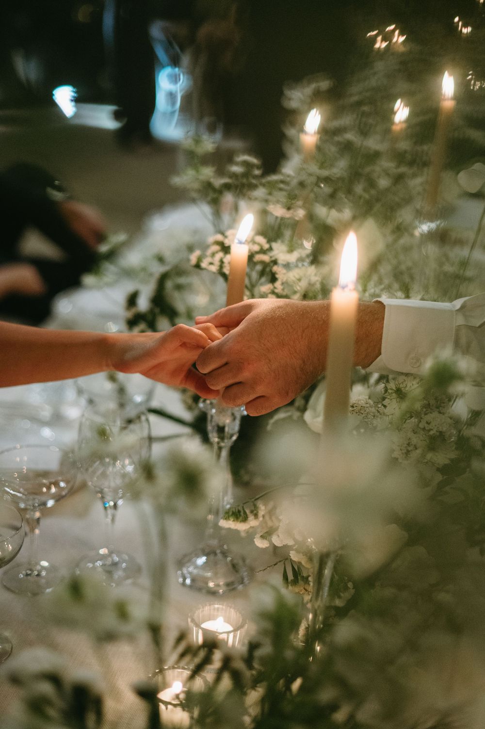 Two hands reach out to touch each other across an elegant table setting with candlelight and floral decorations.