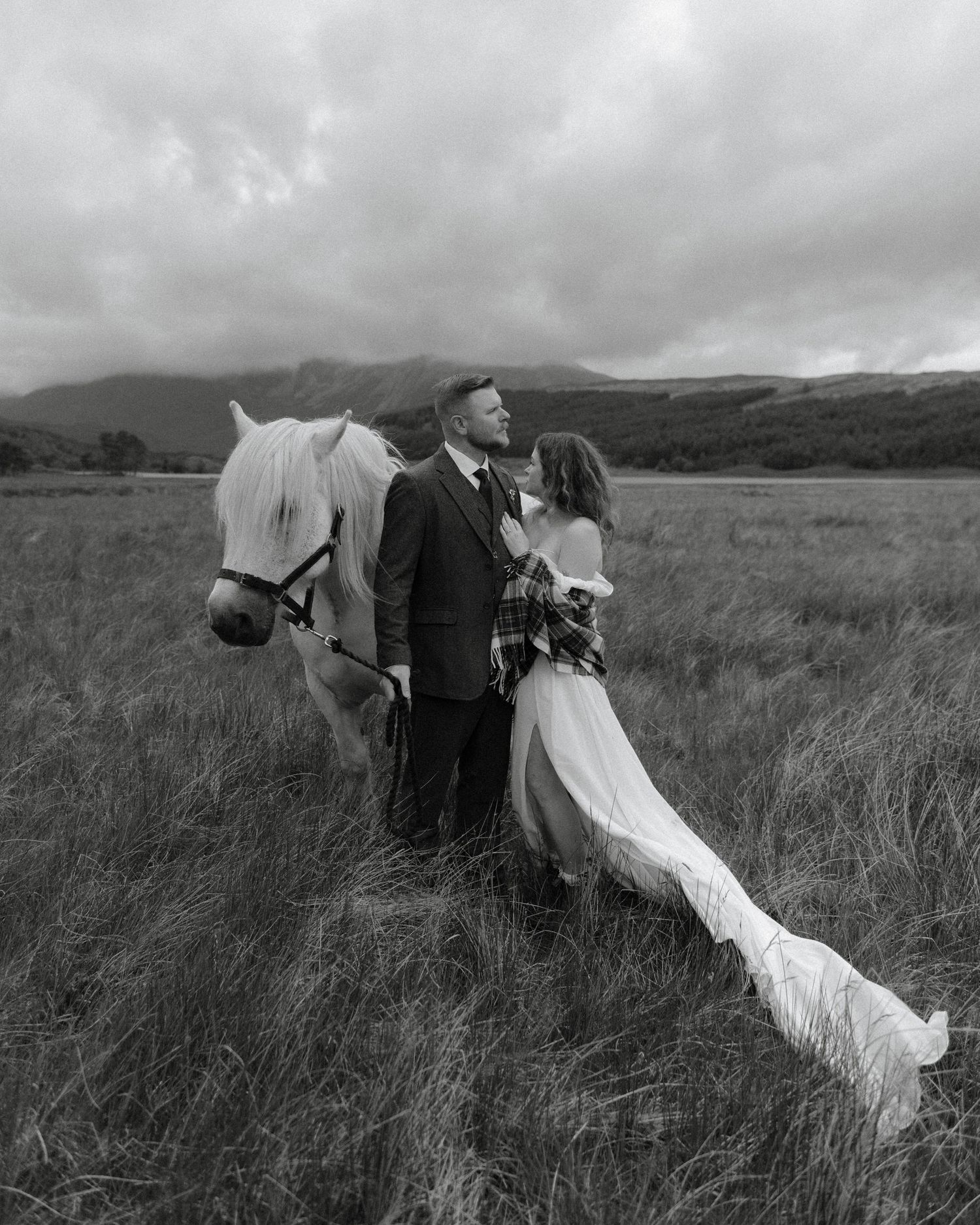 A romantic black and white wedding photo featuring a couple and a white horse in a misty mountain meadow.