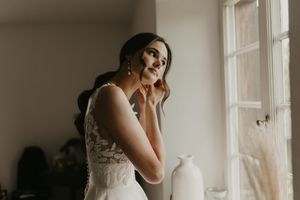 Bride in white dress preparing by window in soft natural light.