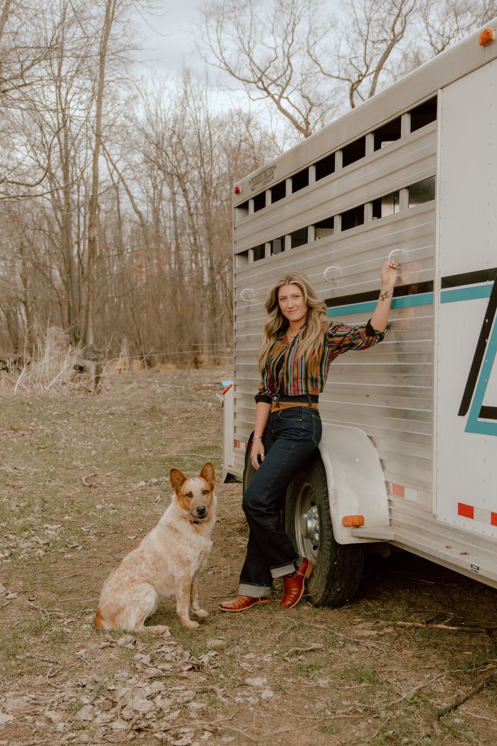 Woman in colorful striped shirt standing beside a horse trailer with a red cattle dog.