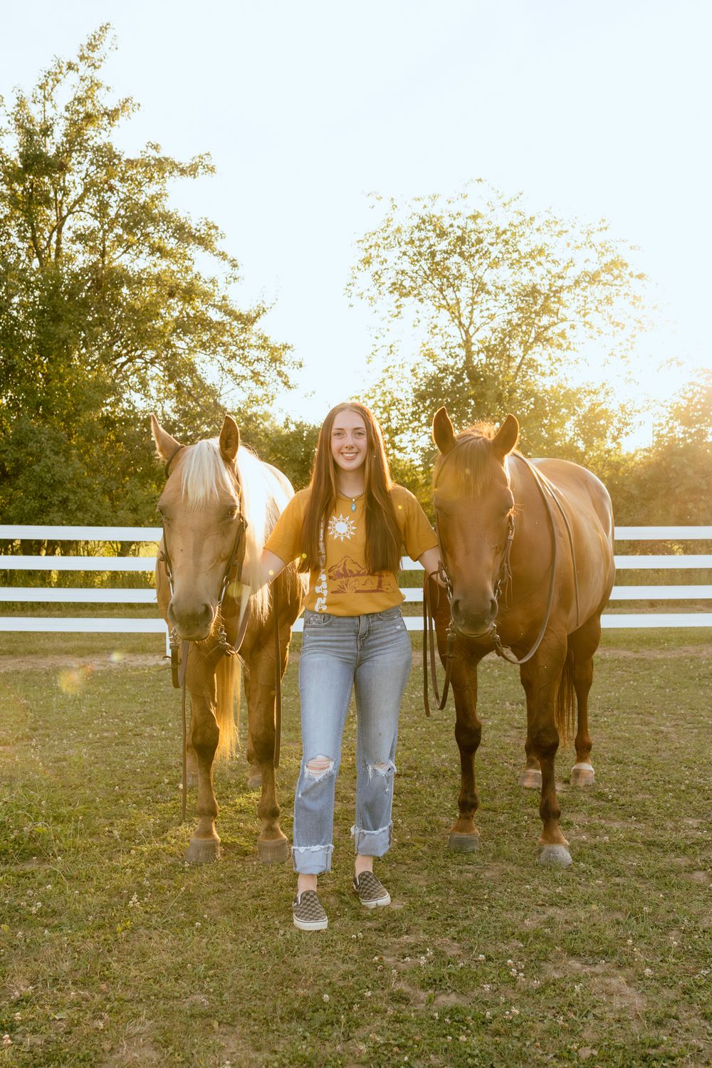 A smiling young woman in jeans and yellow top stands between two brown horses at a sunny white-fenced farm.
