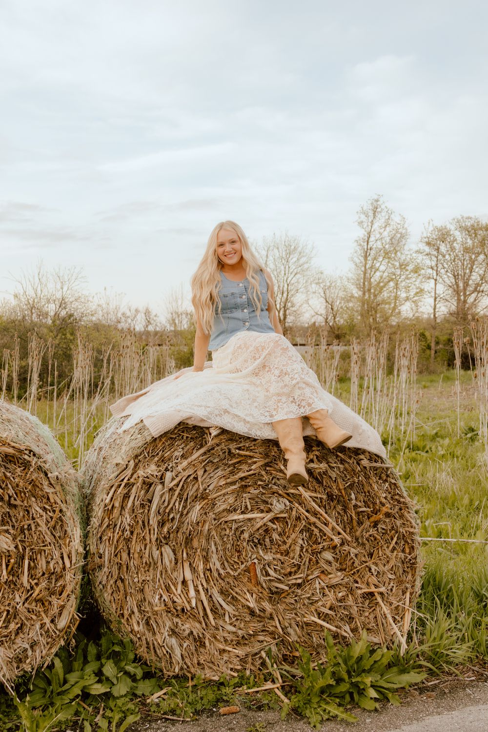 Smiling blonde woman sitting on a large round hay bale in a rural field wearing a denim top and lace skirt.