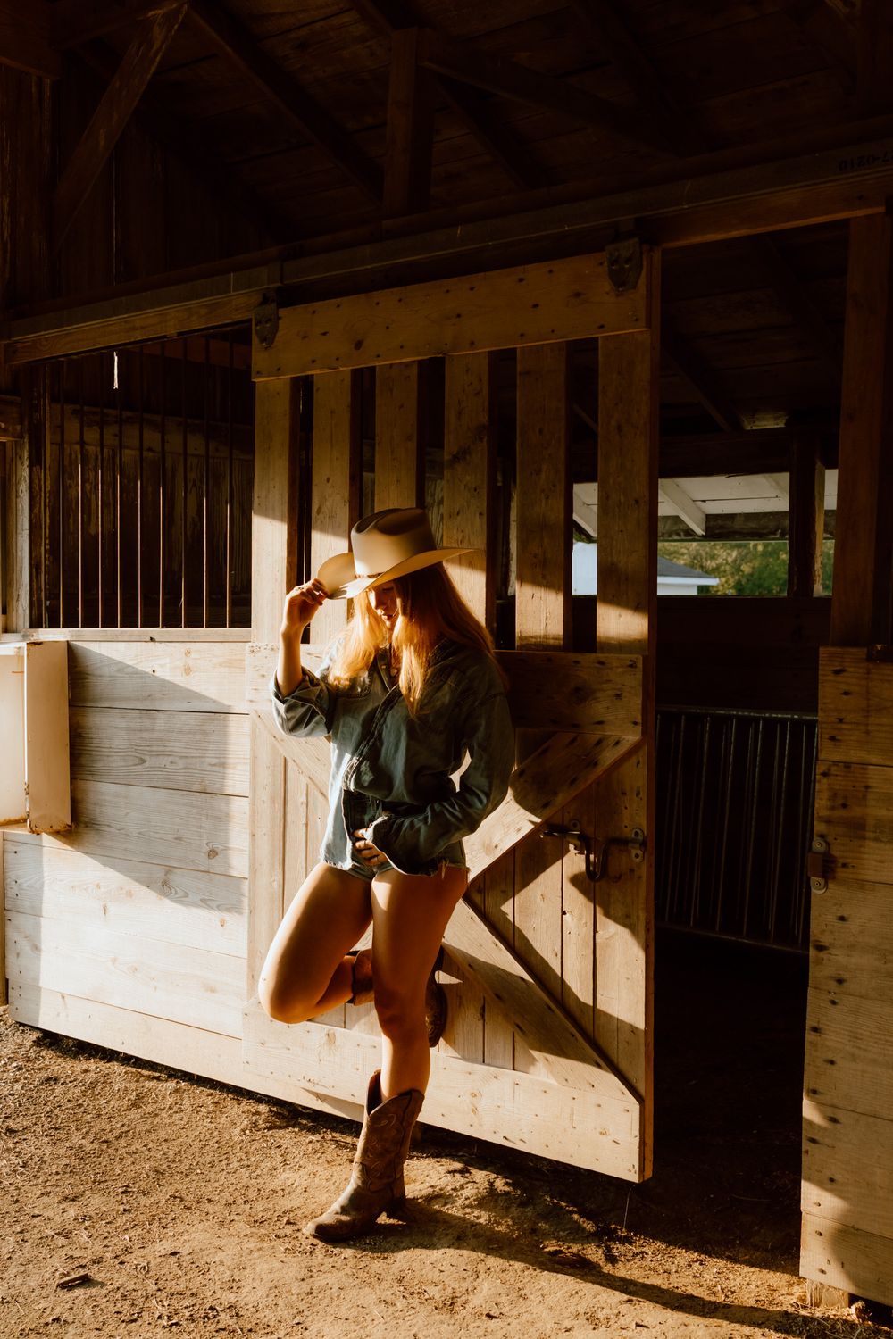 Woman in cowboy hat and boots posing in rustic wooden horse stable with warm golden sunlight.