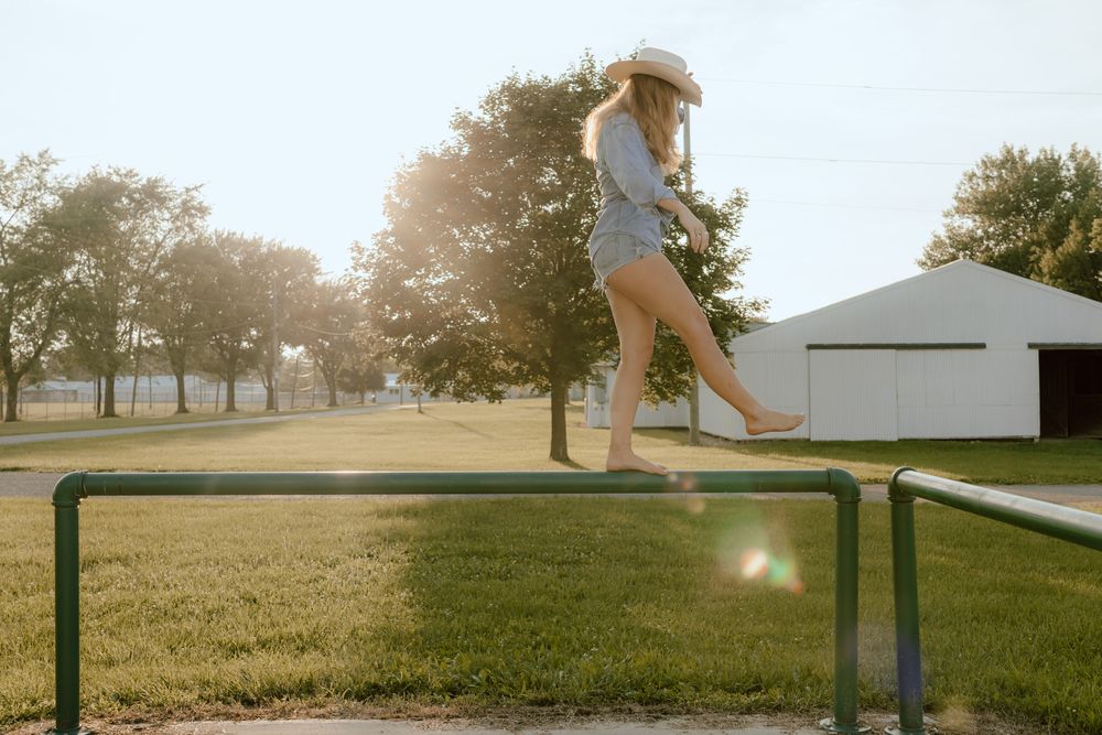 Barefoot blonde woman in cowboy hat balancing on green metal rails in sunny farm field.