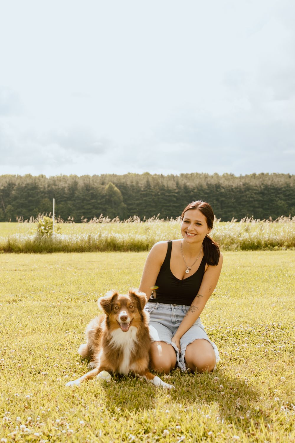 Smiling young woman in black tank top and denim shorts sitting on grass with her Australian Shepherd dog outdoors.