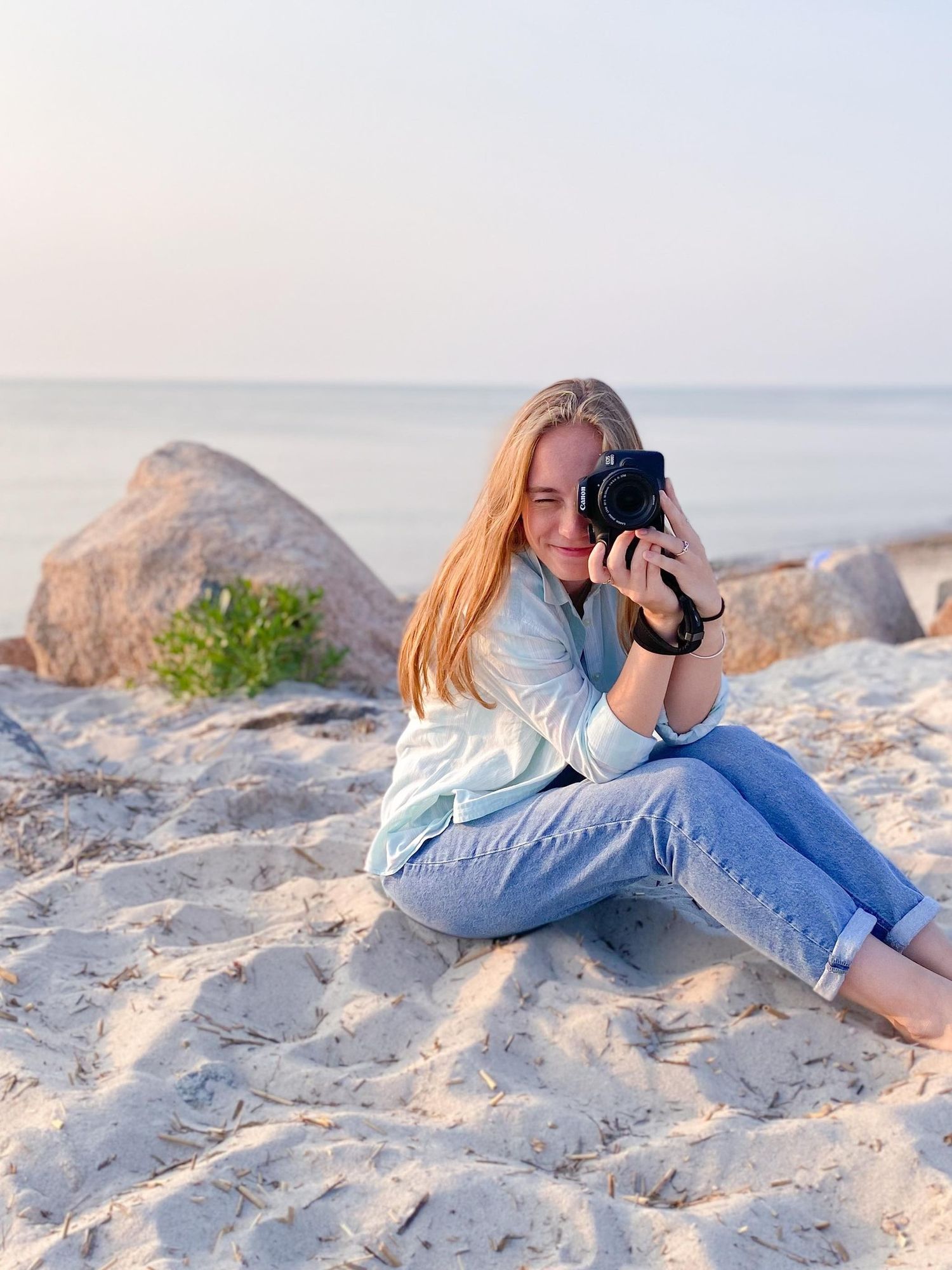 Photographer in a button down shirt and jeans sitting on sandy beach with rocks taking photos at sunset.
