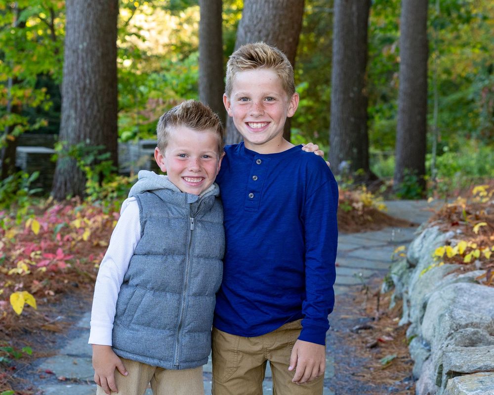Sibling pose for photos in a wooded forest setting with dappled sunlight filtering through trees during family photo session in Sudbury, MA.