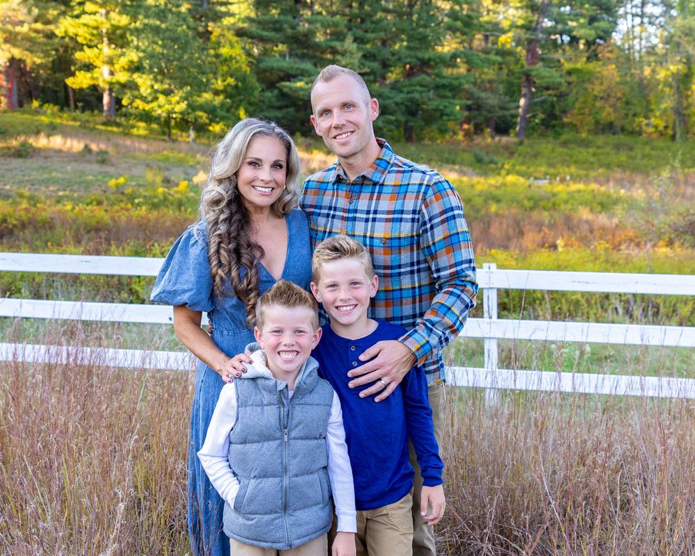 A family poses together in a fall setting with white fences and autumn foliage in the background in coordinated blue outfits during photos.