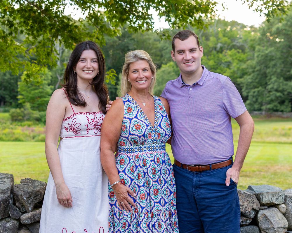 A family in coordinated blue, red & white outfits pose together outdoors near a stone wall at the Codman Estate in Lincoln, MA.