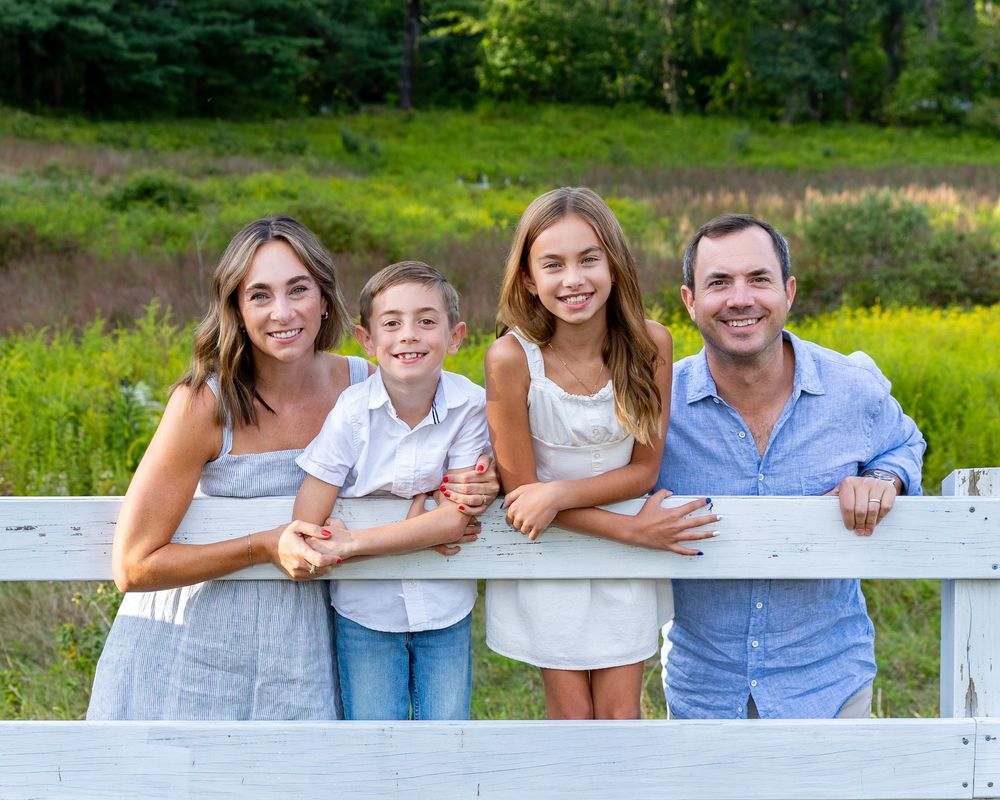 A happy family leaning on a white fence outdoors with green meadow and trees in the background. during their family photo session.
