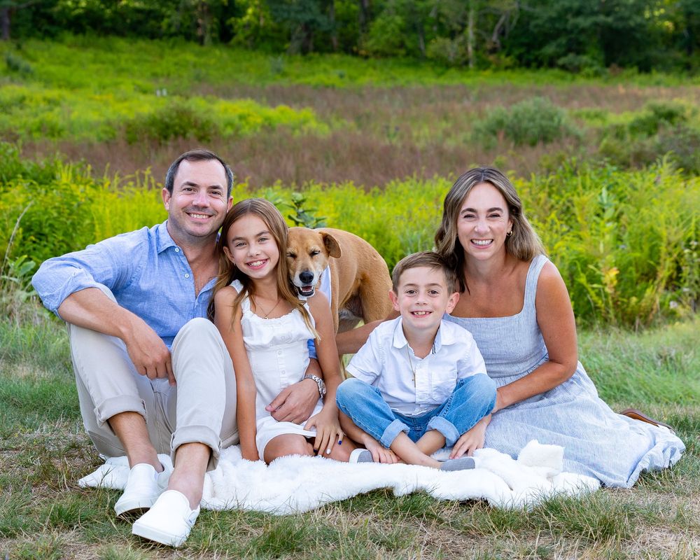 Family and their dog wearing coordinated white outfits sit together on green grass during outdoor photo session.