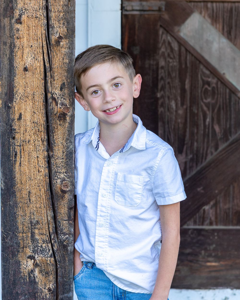 A young boy in a white button-down shirt and jeans stands smiling against a rustic wooden wall and post in portrait during family photos.