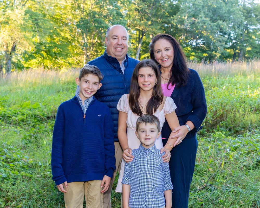 A family poses together outdoors in coordinated blue and white outfits against a natural backdrop of trees and grass.